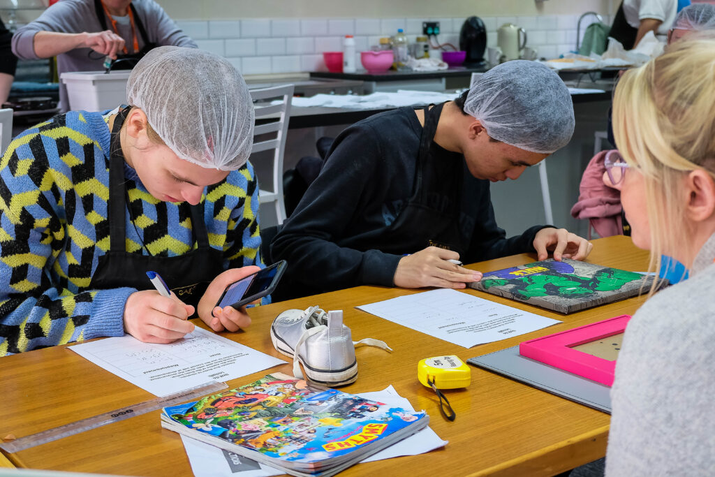 One female and one male learner, sat at a desk wearing aprons and hair nets. They are working on an arts and crafts project with colourful materials and a host of tools, including a mobile device