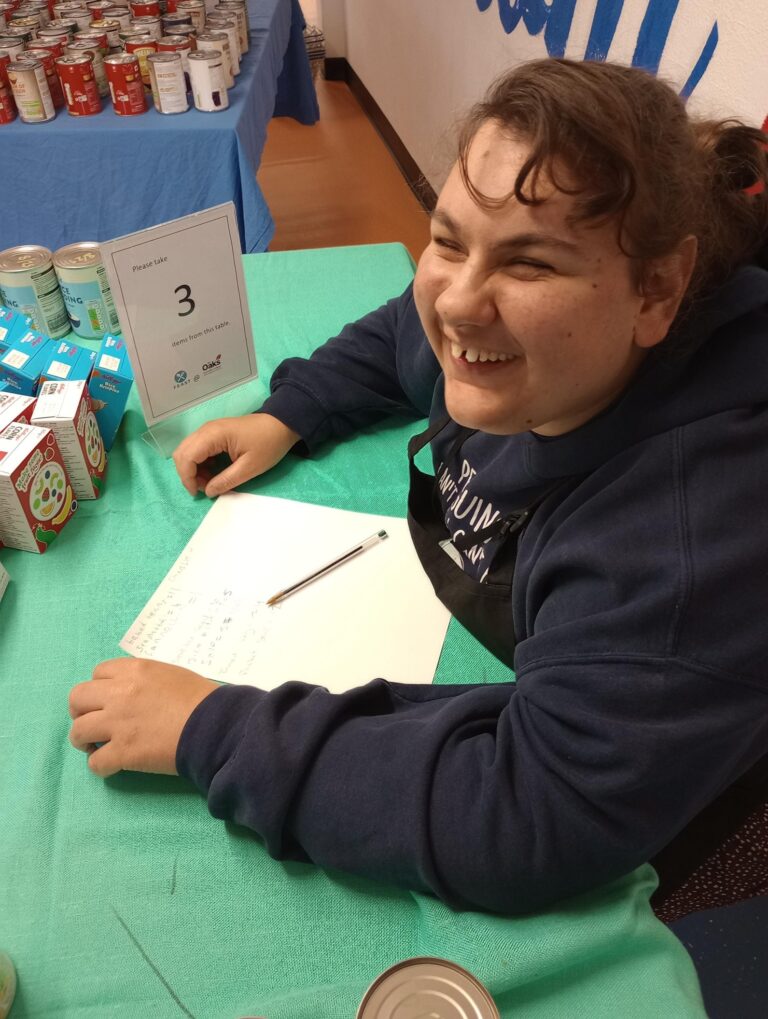 A learner, smiling broadly at the welcome desk of the college's community food larder, run with F.E.A.S.T